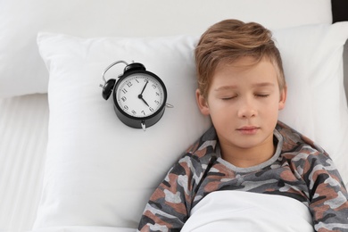 Cute boy with alarm clock sleeping in bed, above view