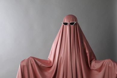 Glamorous ghost. Woman in pink sheet with sunglasses on light grey background, space for text