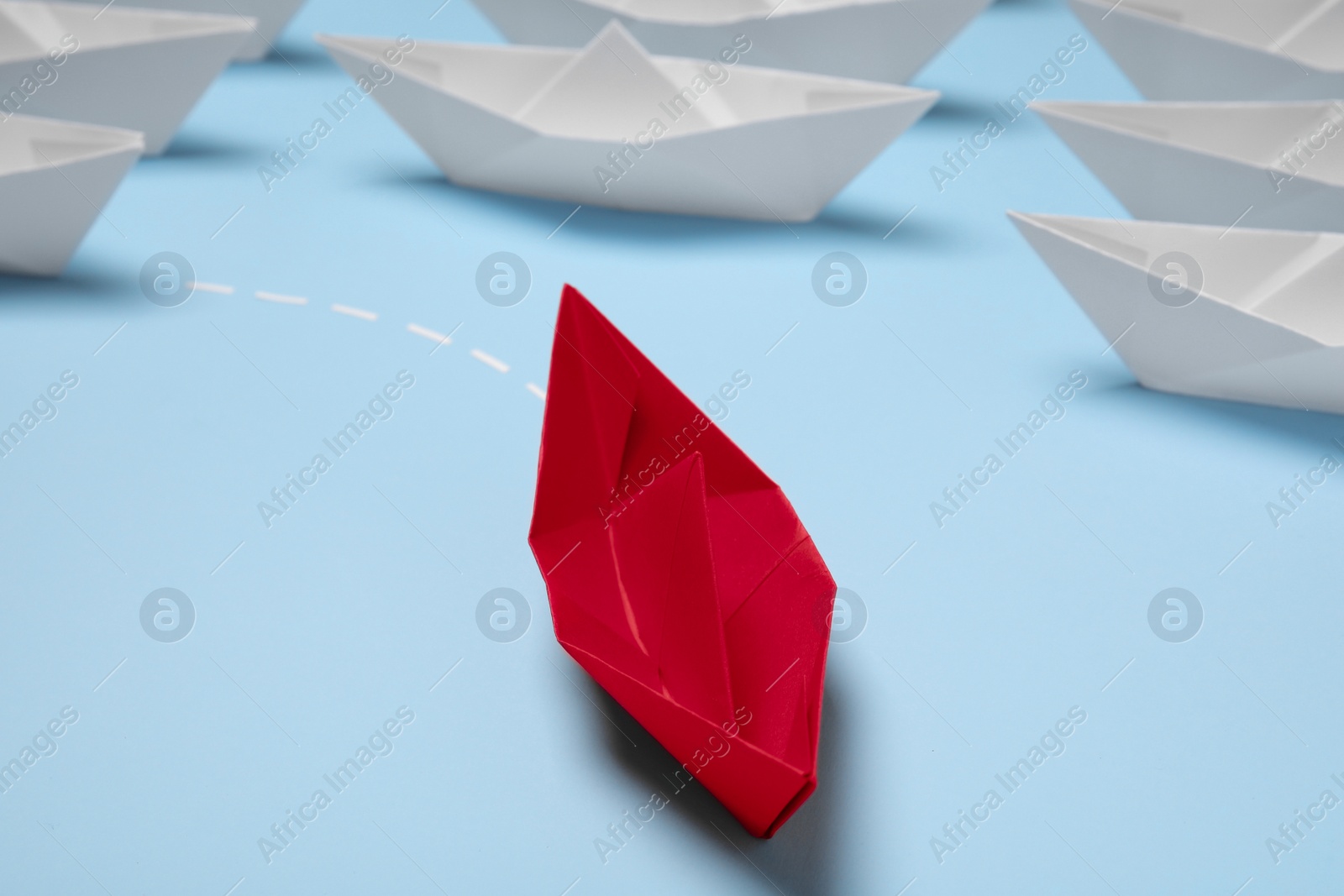 Photo of Red paper boat floating away from others on light blue background, closeup. Uniqueness concept