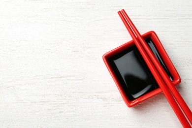 Dish of soy sauce with chopsticks on white wooden background, flat lay. Space for text