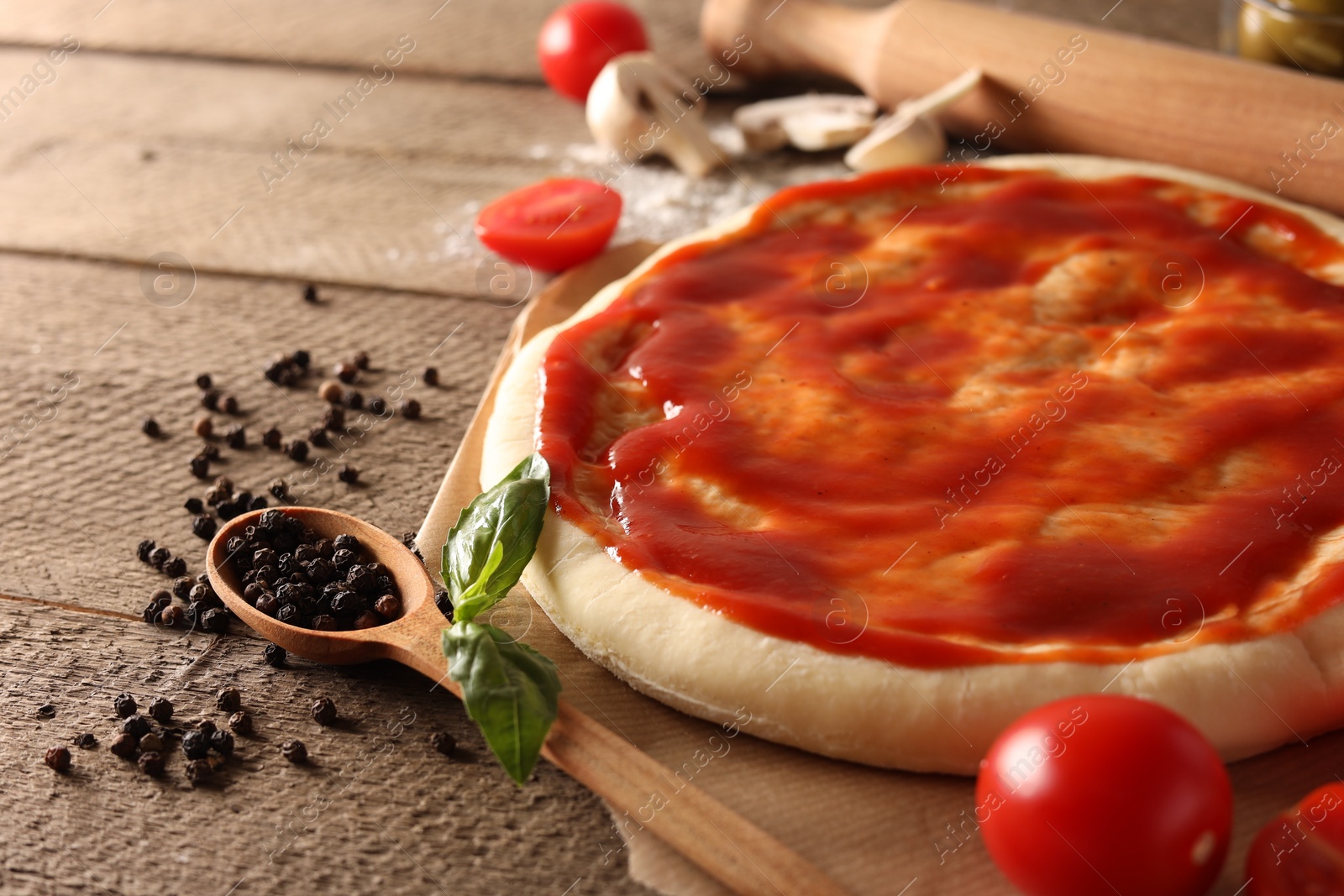 Photo of Pizza dough smeared with tomato sauce and products on wooden table, closeup