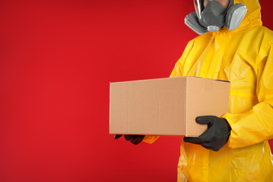 Man in chemical protective suit holding cardboard box on red background, closeup view with space for text. Prevention of virus spread