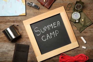 Photo of Chalkboard with text SUMMER CAMP and camping equipment on wooden background, flat lay