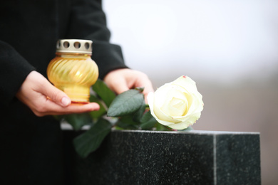 Photo of Woman with candle and rose near black granite tombstone outdoors, closeup. Funeral ceremony
