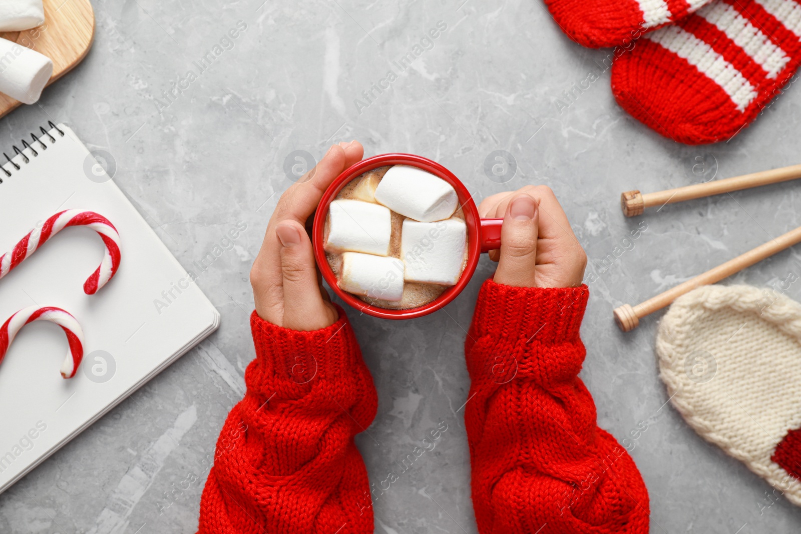 Photo of Woman holding cup of tasty cocoa with marshmallows at grey marble table, top view. Cozy winter