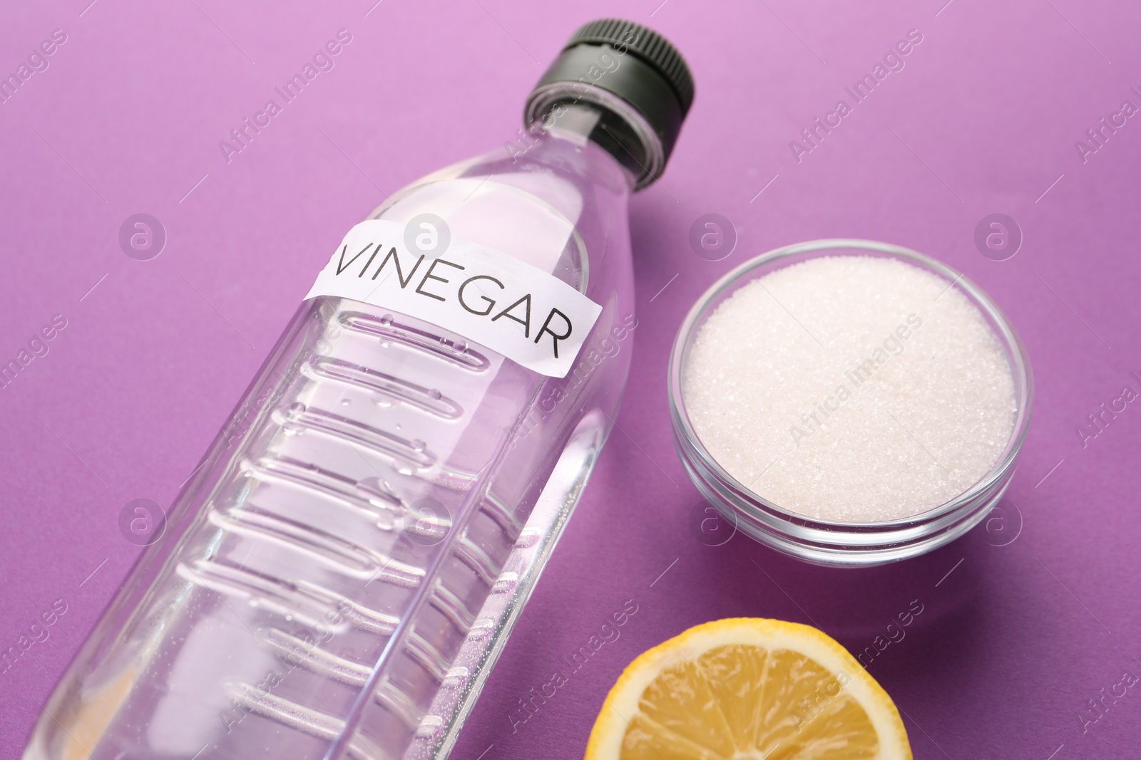 Photo of Eco friendly natural cleaners. Vinegar in bottle, cut lemon and bowl of soda on purple background