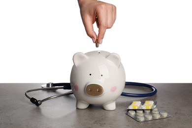 Photo of Woman putting coins into piggy bank on grey table against white background, closeup. Medical insurance