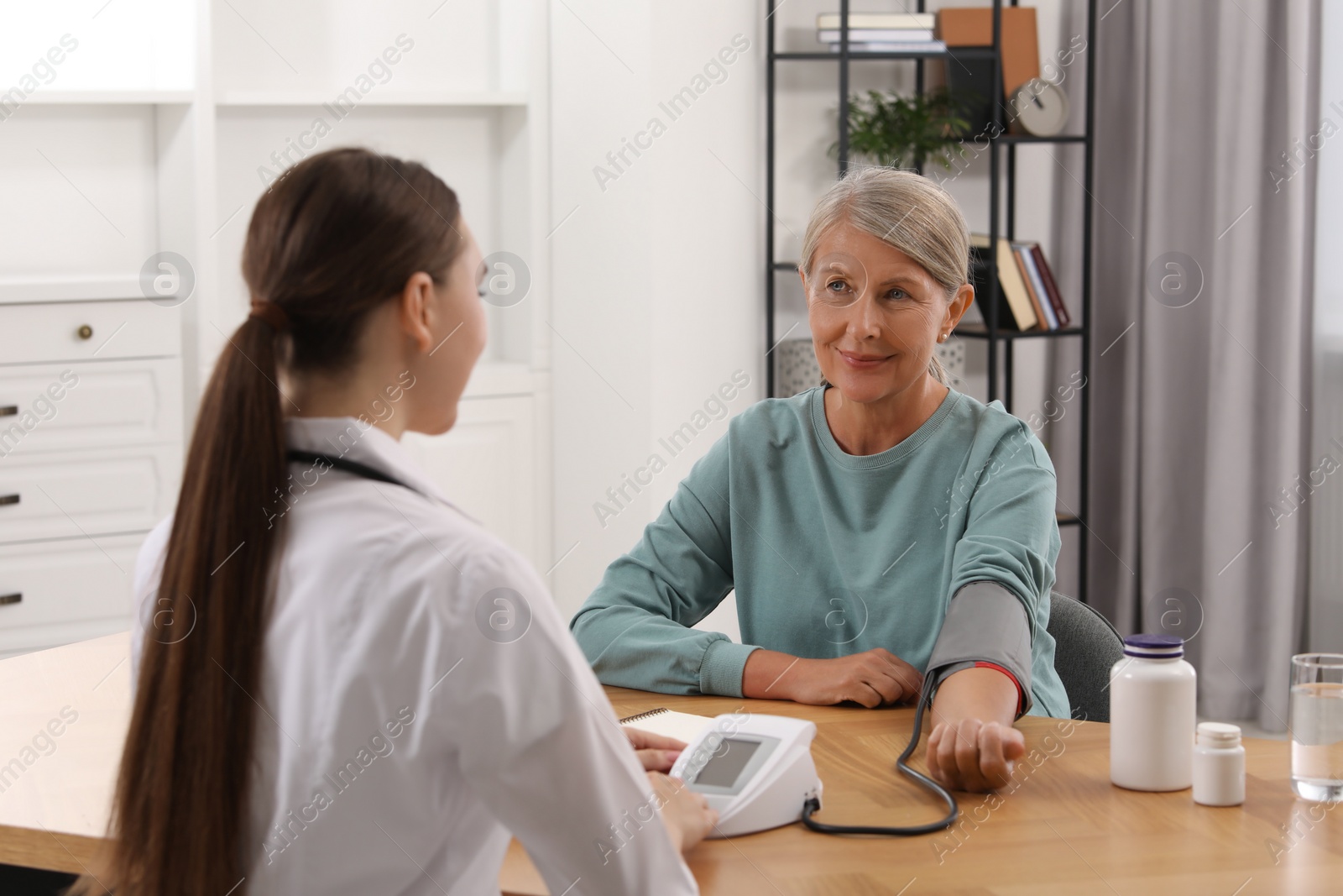 Photo of Young healthcare worker measuring senior woman's blood pressure at wooden table indoors