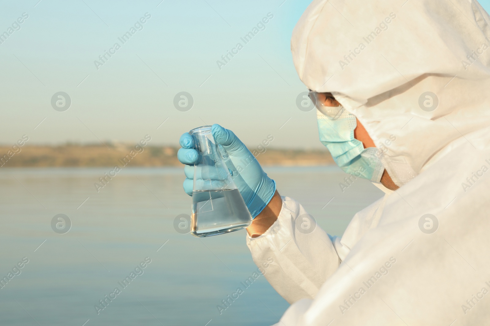 Photo of Scientist in chemical protective suit with conical flask taking sample from river for analysis