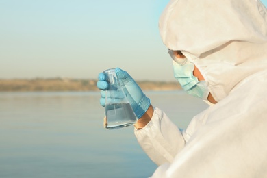 Photo of Scientist in chemical protective suit with conical flask taking sample from river for analysis