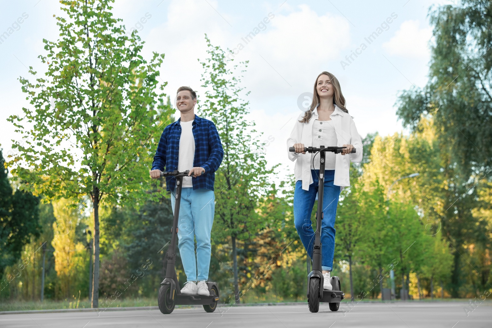 Photo of Happy couple riding modern electric kick scooters in park, low angle view