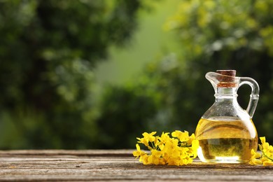 Photo of Rapeseed oil in glass jug and beautiful yellow flowers on wooden table outdoors, space for text