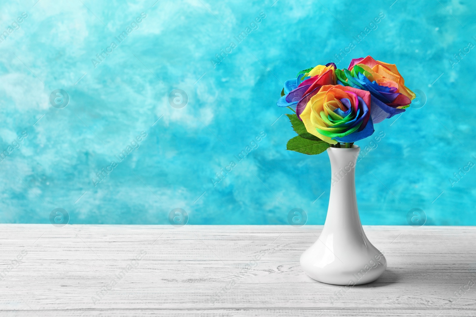 Photo of Vase with rainbow rose flowers on table against color background