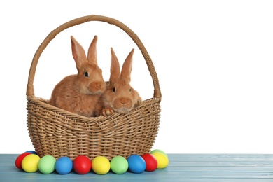 Cute fluffy bunnies in wicker basket near Easter eggs on light blue wooden table. Space for text