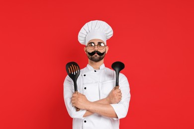 Photo of Professional chef with funny artificial moustache holding kitchen utensils on red background