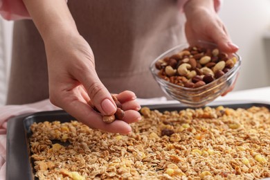 Photo of Making granola. Woman adding nuts onto baking tray with mixture of oat flakes and other ingredients at table in kitchen, closeup