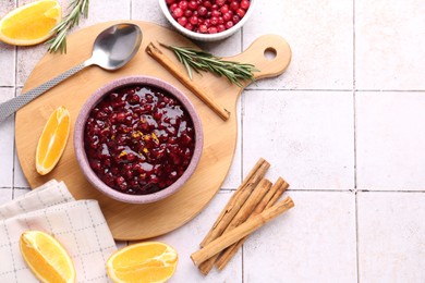 Photo of Tasty cranberry sauce in bowl and ingredients on white tiled table, flat lay. Space for text