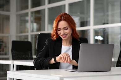 Photo of Happy woman checking time while working with laptop at white desk in office