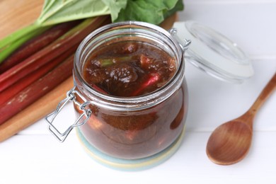 Jar of tasty rhubarb jam, stems and spoon on white table, closeup