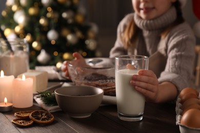 Photo of Little child making Christmas pastry at wooden table, closeup. Focus on glass of milk
