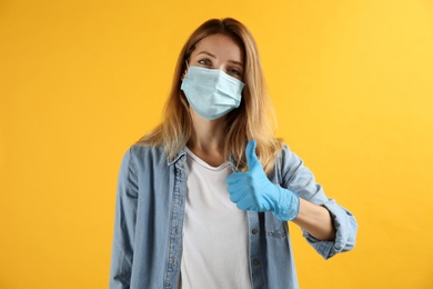 Photo of Young woman in medical gloves and protective mask showing thumb up on yellow background