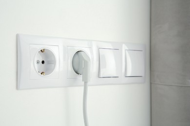 Power sockets with inserted plug and light switches on white wall indoors