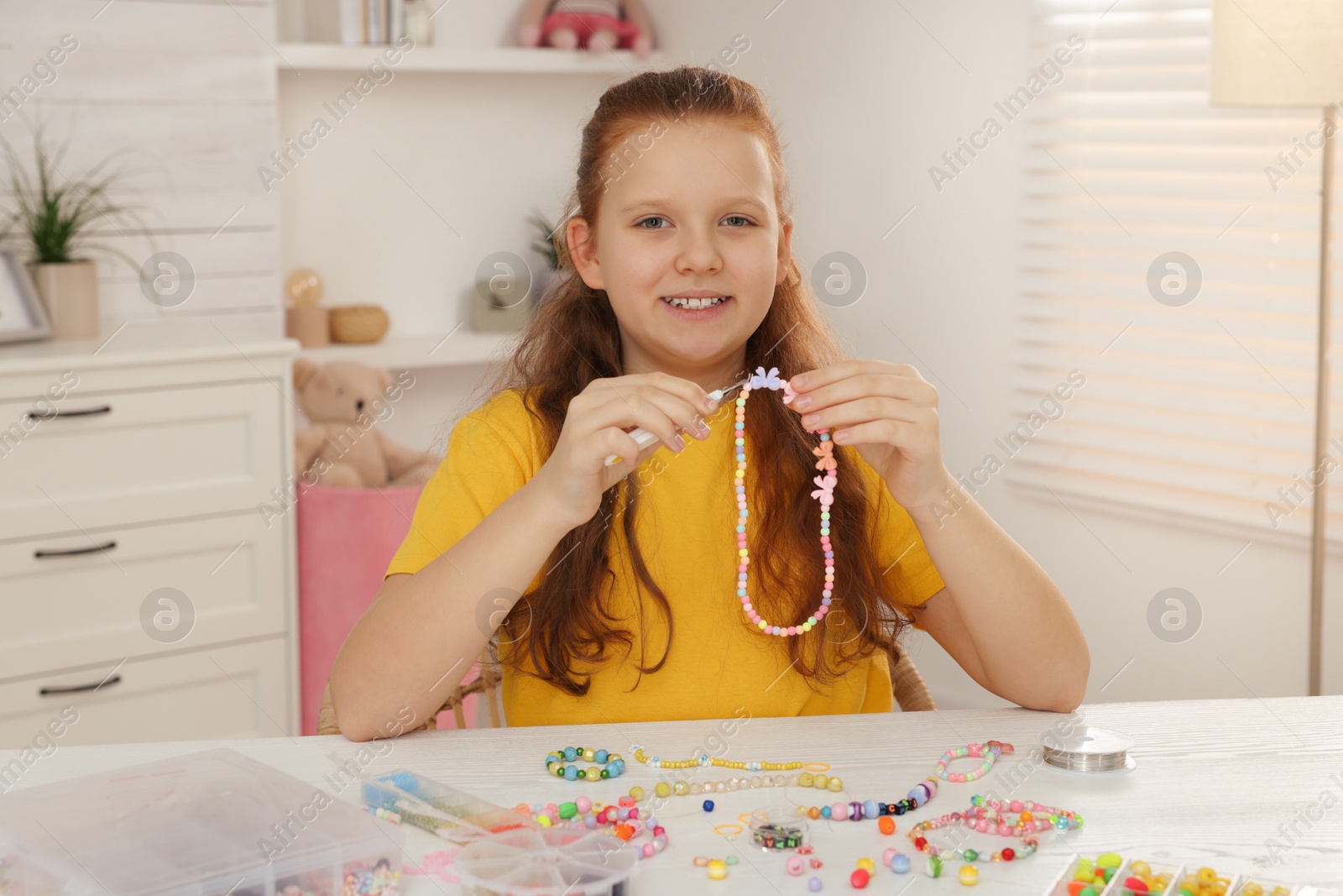 Photo of Cute girl making beaded jewelry at table in room