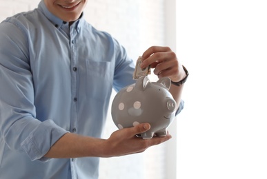 Photo of Young businessman putting money into piggy bank indoors, closeup. Space for text