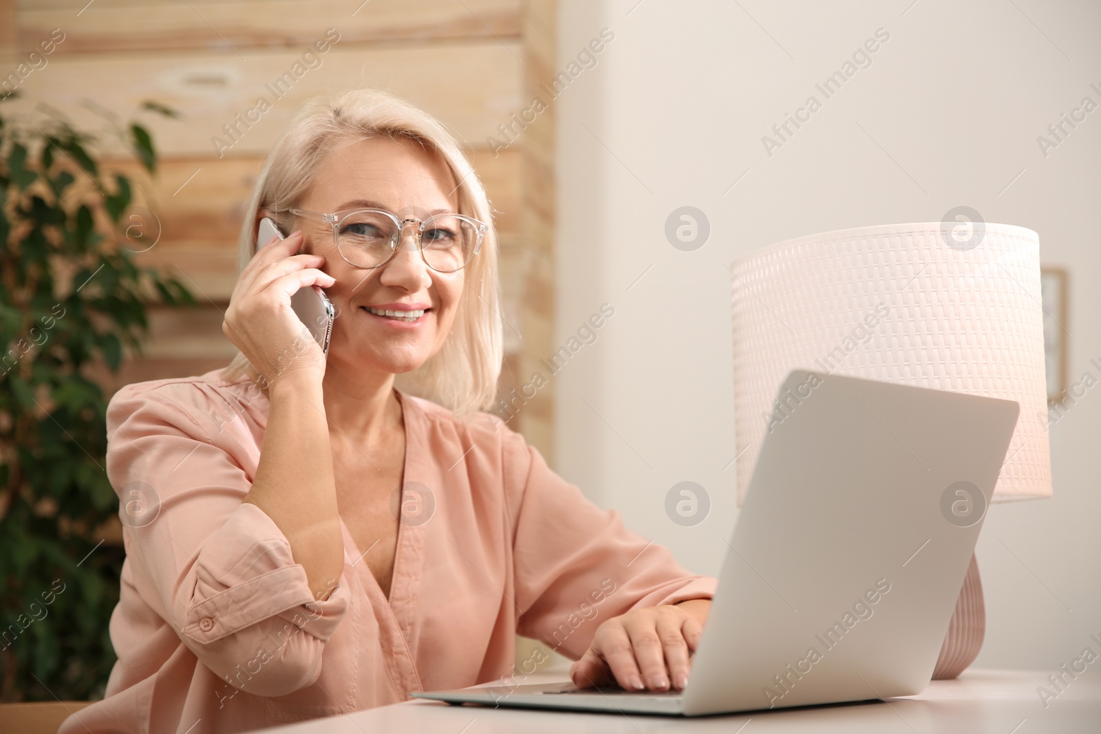 Photo of Beautiful mature woman with laptop talking on smartphone at home