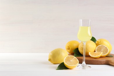 Liqueur glass with tasty limoncello, lemons and green leaves on white wooden table. Space for text