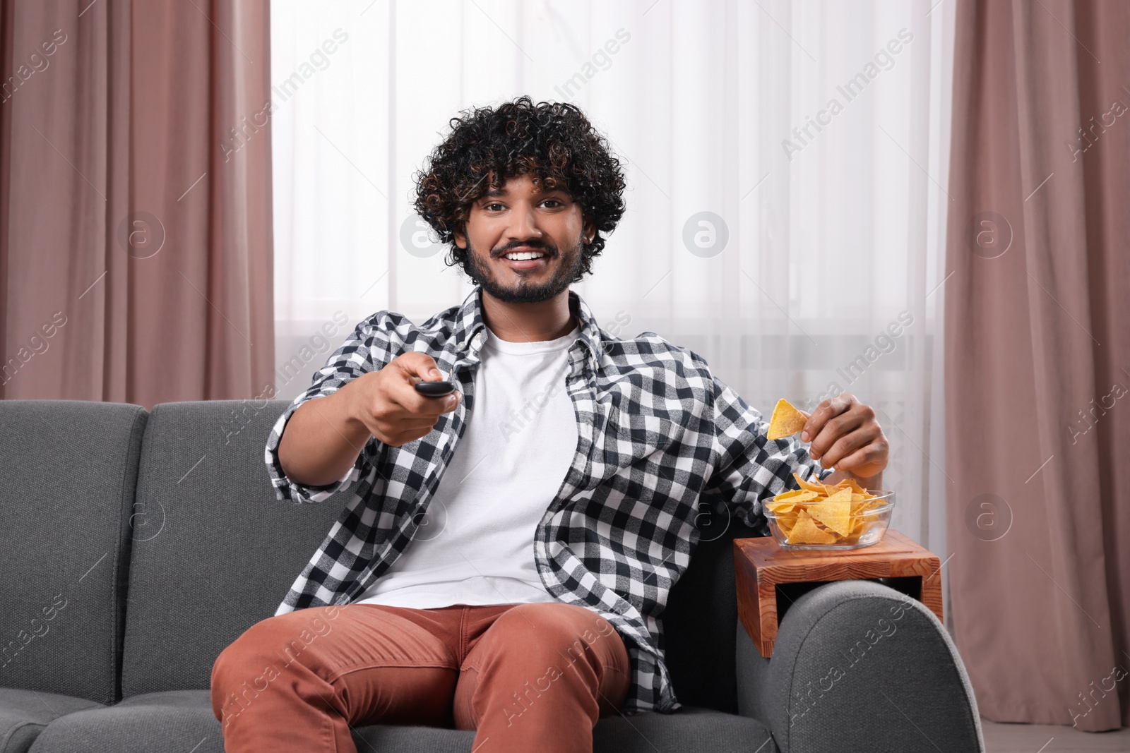 Photo of Happy man switching TV channels and eating nacho chips on sofa with wooden armrest table at home