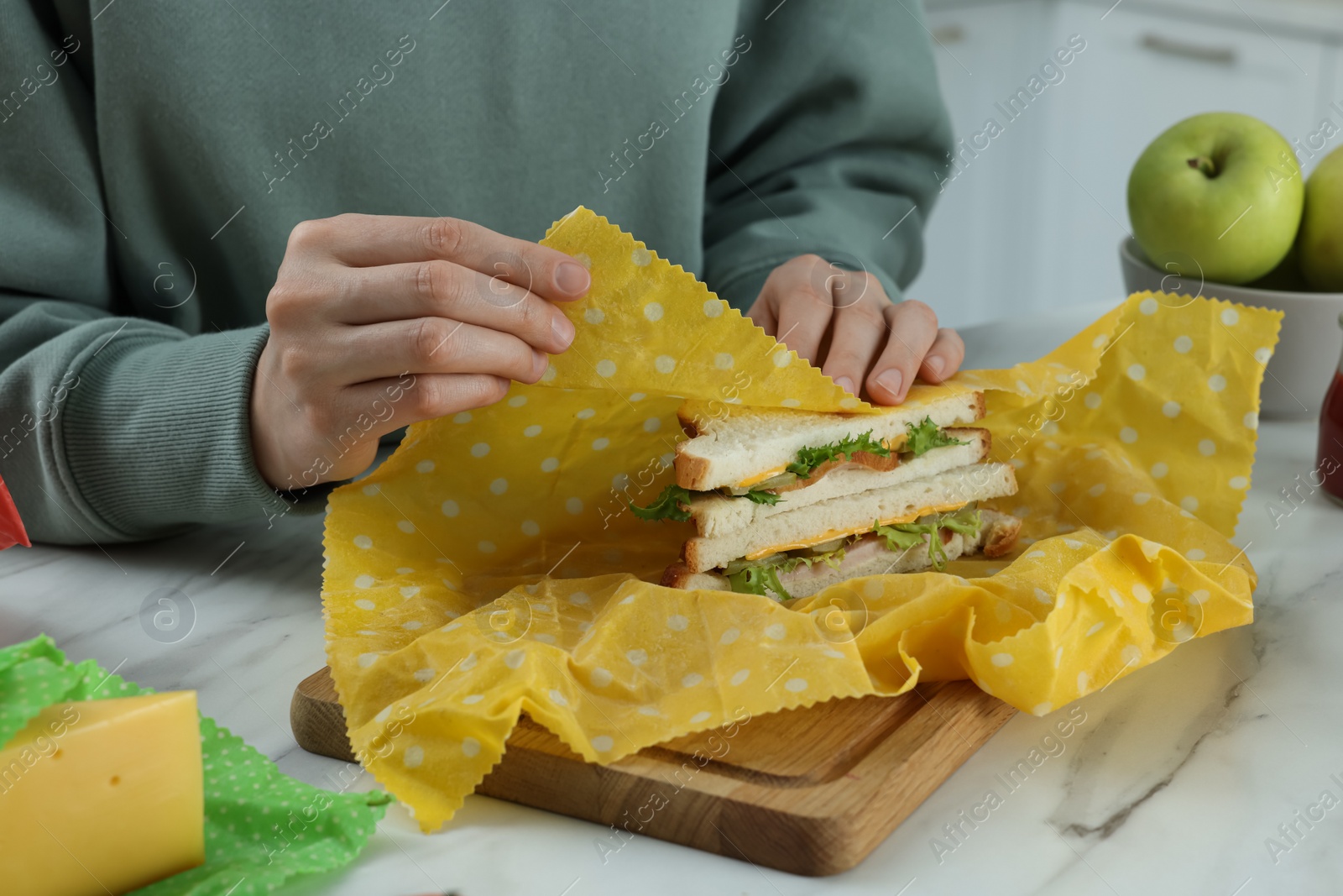 Photo of Woman packing tasty sandwich into beeswax food wrap at white marble table indoors, closeup