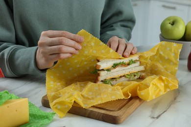 Photo of Woman packing tasty sandwich into beeswax food wrap at white marble table indoors, closeup