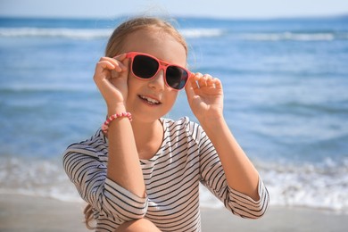 Photo of Happy little girl in stylish sunglasses on beach near sea