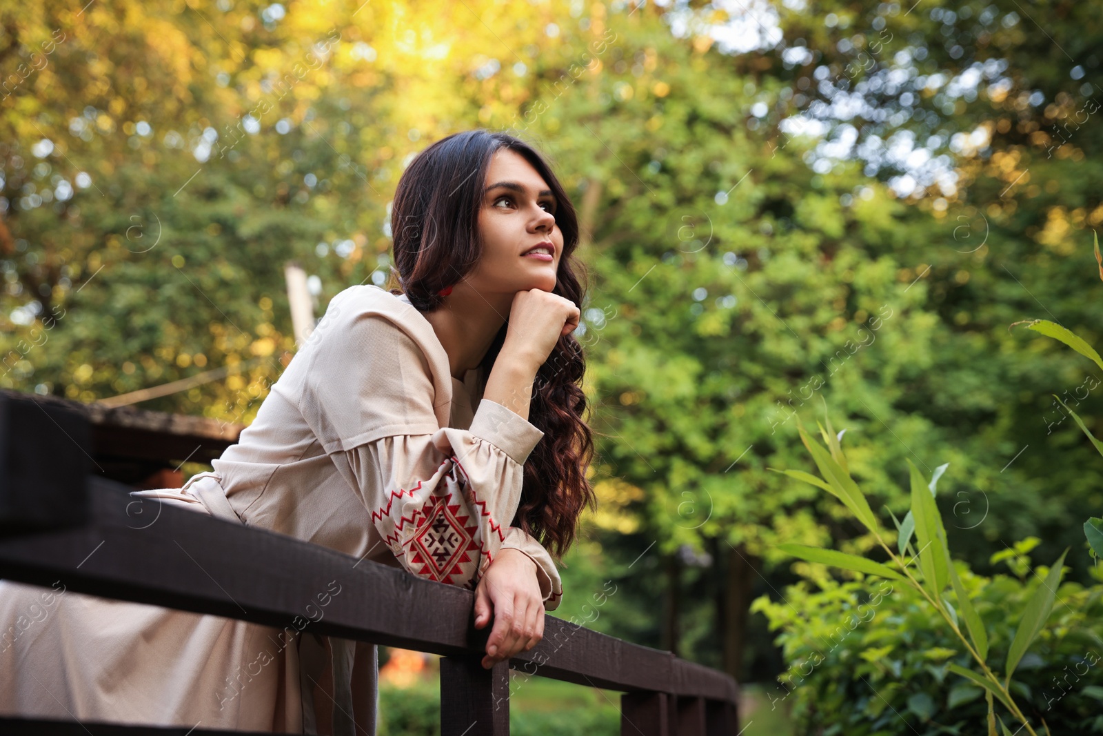 Photo of Beautiful woman wearing embroidered dress near wooden railing in countryside, space for text. Ukrainian national clothes