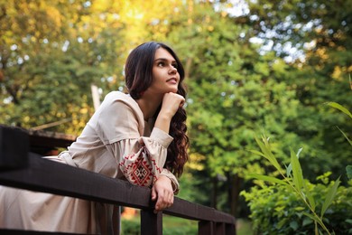 Photo of Beautiful woman wearing embroidered dress near wooden railing in countryside, space for text. Ukrainian national clothes