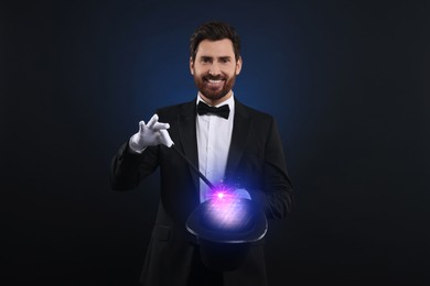 Magician showing trick with wand and top hat on dark background. Fantastic light coming out of hat