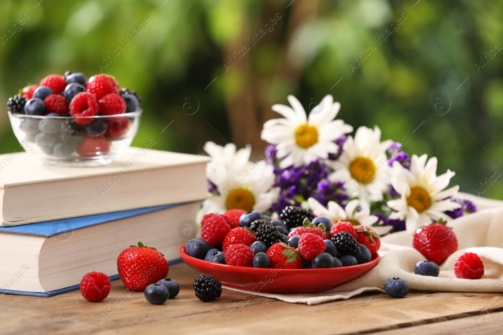 Photo of Different fresh ripe berries, beautiful flowers and books on wooden table outdoors