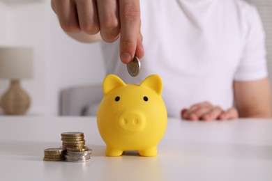 Man putting coin into yellow piggy bank at white table, closeup