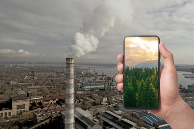 Environmental pollution. Man holding phone with beautiful mountain landscape on screen against industrial factory