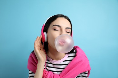 Photo of Fashionable young woman with headphones blowing bubblegum on light blue background