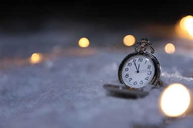 Photo of Pocket watch and Christmas lights on white snow outdoors, space for text. Midnight countdown