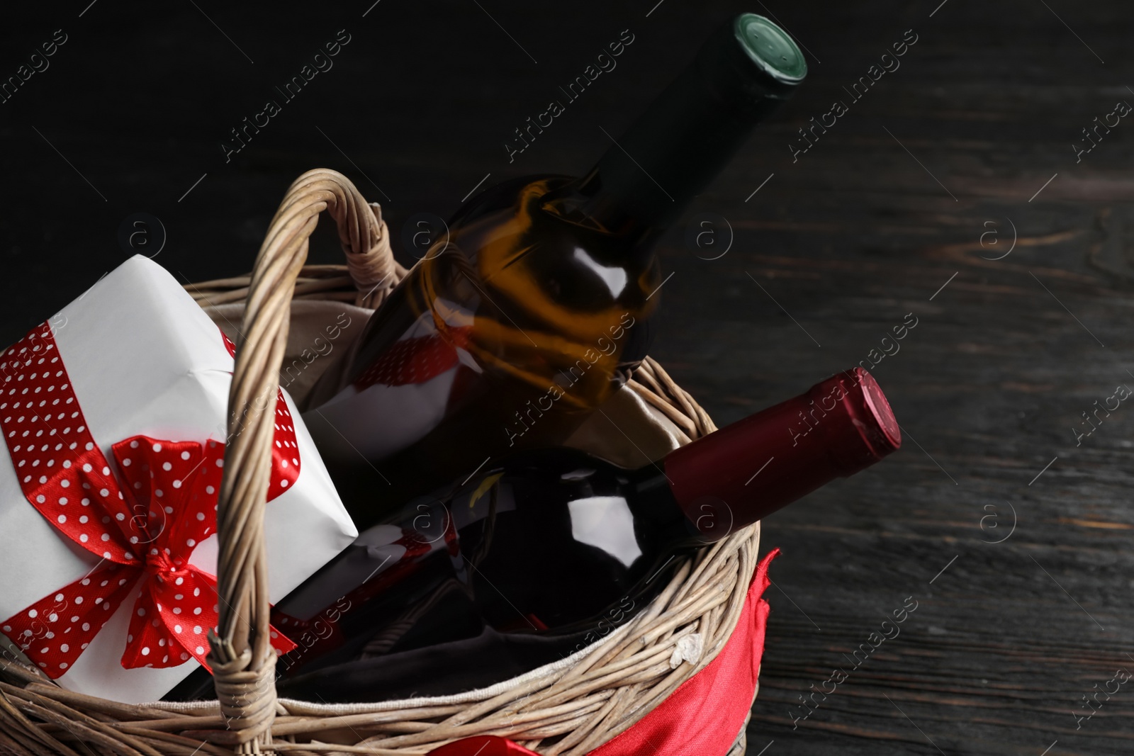 Photo of Festive basket with bottles of wine and gift on dark table, closeup