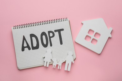 Family figure, house and notebook with word Adopt on pink background, flat lay