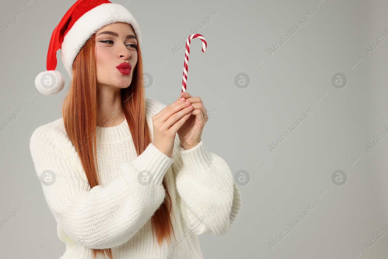 Photo of Young woman in Santa hat with candy cane on light grey background, space for text. Christmas celebration