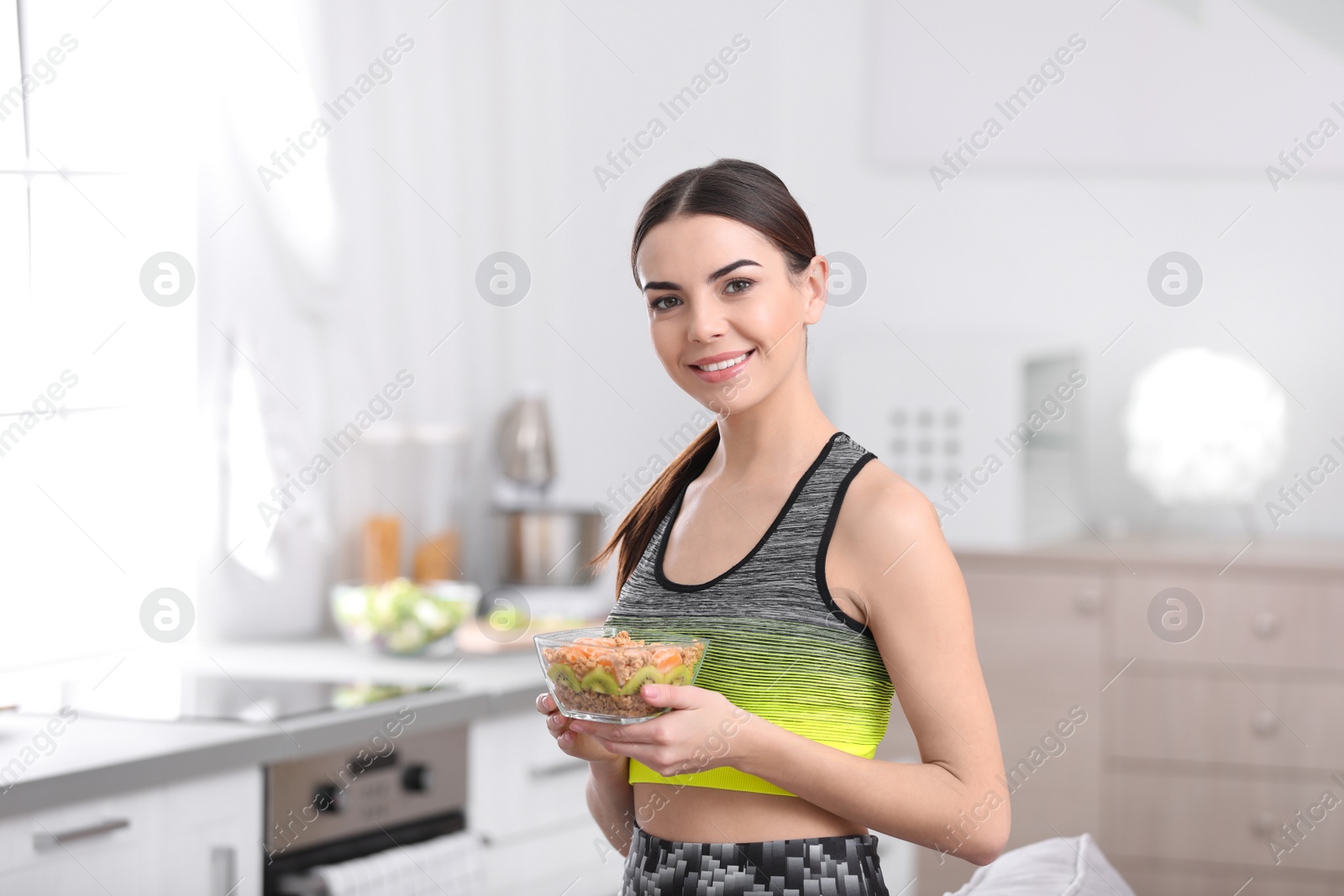 Photo of Young woman in fitness clothes holding bowl of cereal breakfast with fruits at home