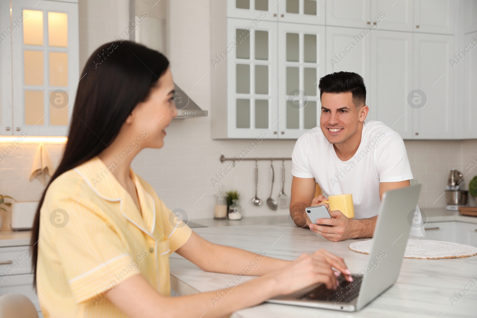 Photo of Happy couple wearing pyjamas with gadgets spending time together in kitchen