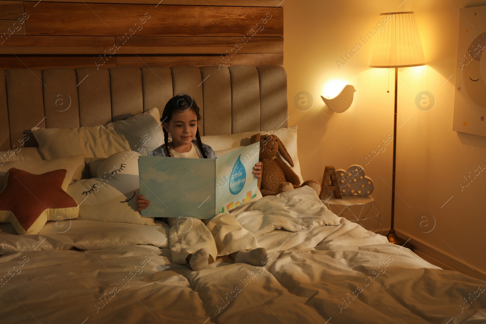 Photo of Little girl reading book in bedroom lit by night lamp