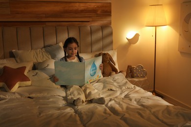 Photo of Little girl reading book in bedroom lit by night lamp
