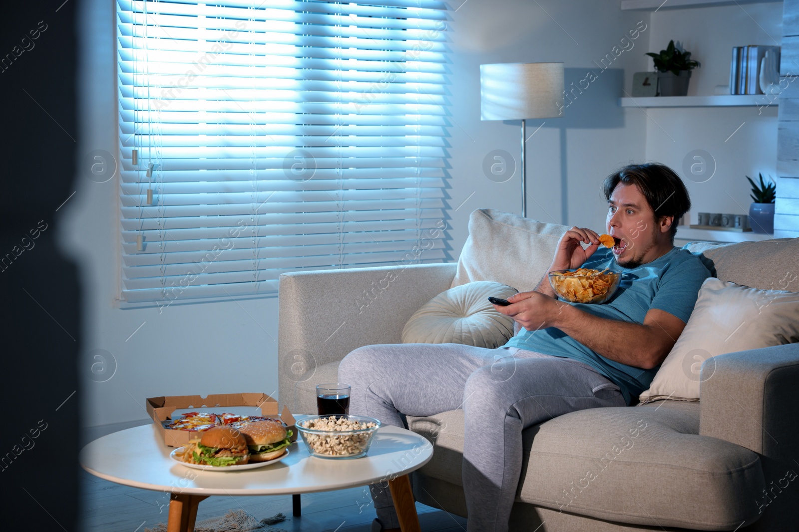 Photo of Overweight man with chips watching TV on sofa at home